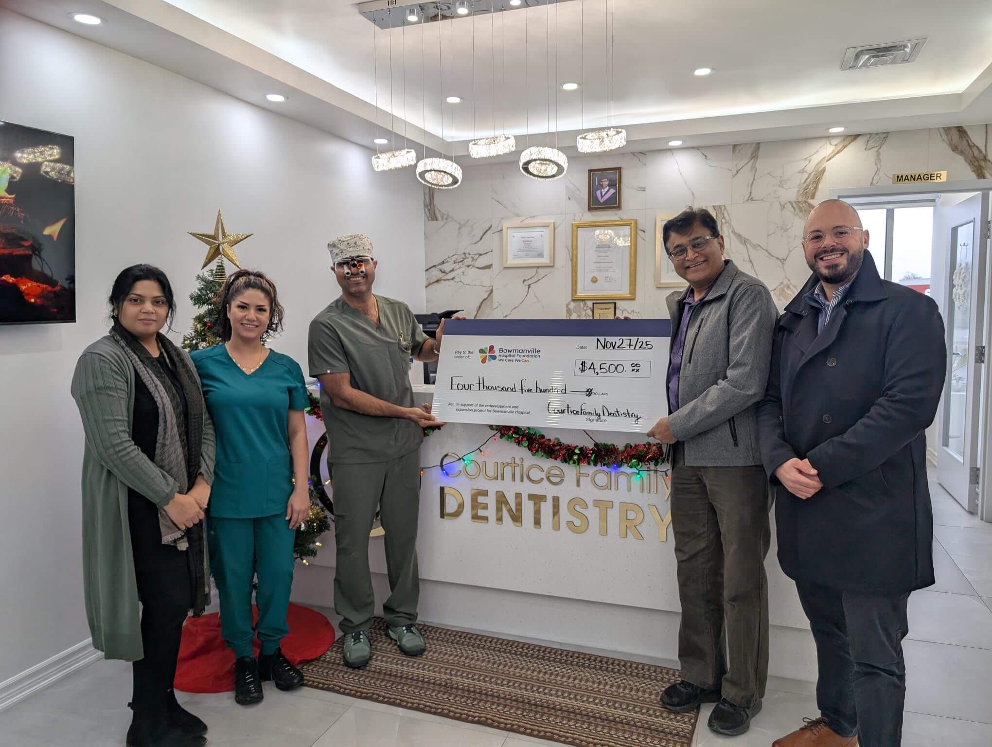 Group of six people standing in a dental office, holding a large donation cheque in front of the reception desk.