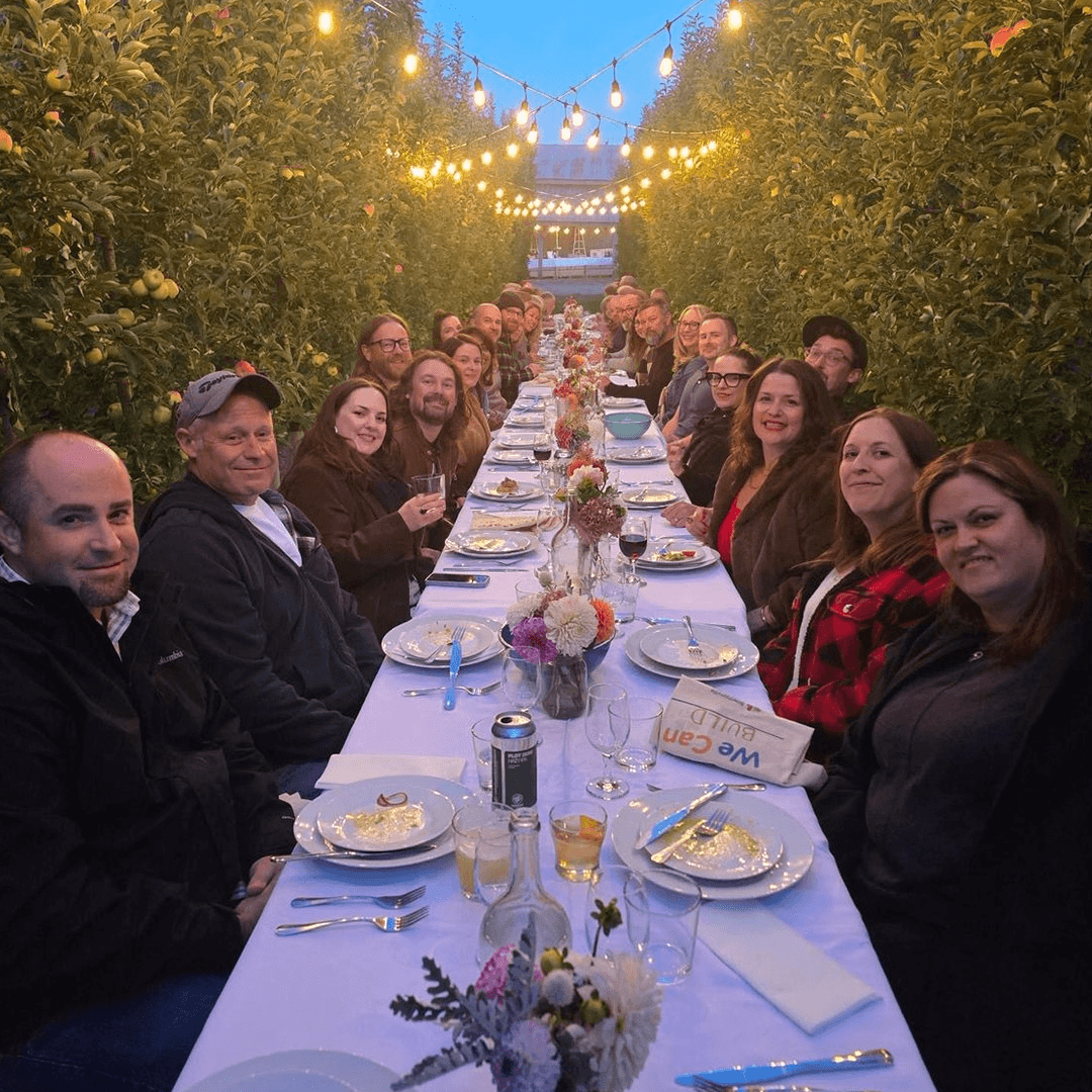 A group of people gathered around a long table in an orchard at dusk.