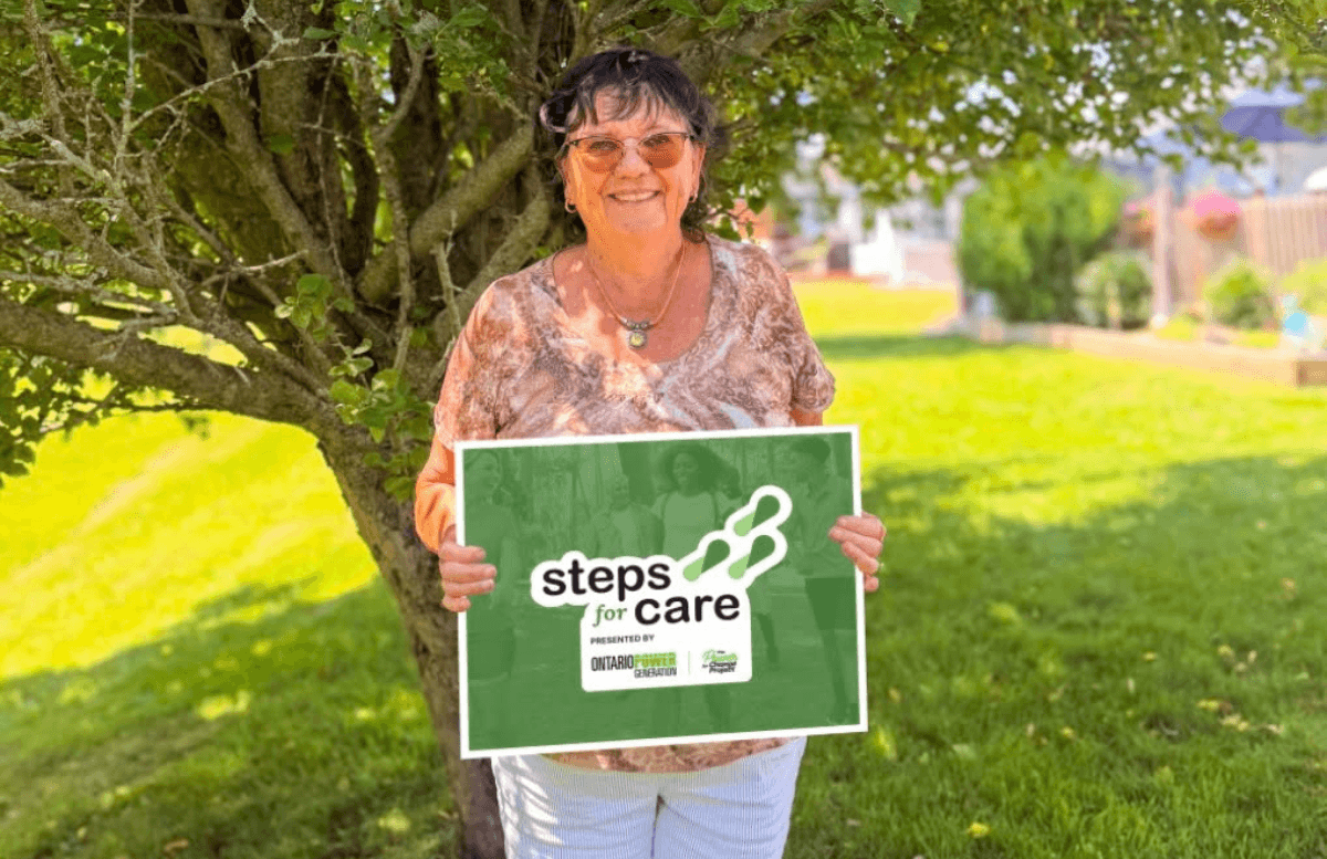 A woman standing underneath a large tree holding a Steps for Care poster.