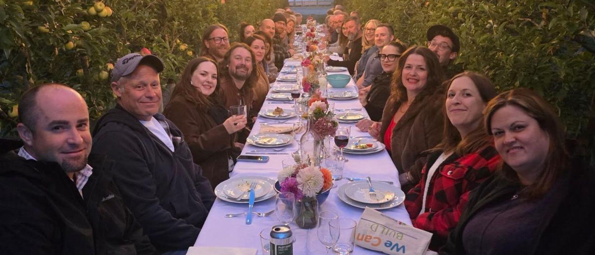 A group of people sitting at a long table in an apple orchard.