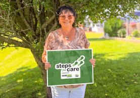 A woman standing underneath a large tree holding a Steps for Care poster.